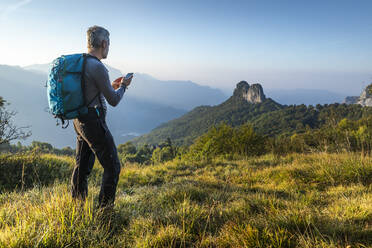 Älterer Mann benutzt sein Smartphone beim Wandern auf einem Berg bei Sonnenaufgang, Orobie, Lecco, Italien - MCVF00591