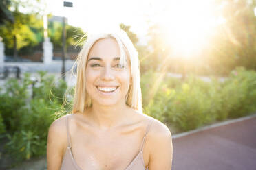 Nahaufnahme einer lächelnden jungen Frau mit blondem Haar im Park bei Sonnenuntergang - DAMF00455