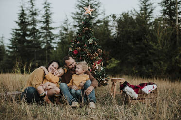 Lächelnde Familie sitzt am Weihnachtsbaum auf einer Wiese auf dem Lande bei Sonnenuntergang - GMLF00603