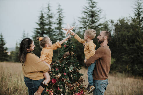 Eltern mit Kindern beim Schmücken des Weihnachtsbaums auf dem Lande bei Sonnenuntergang - GMLF00606