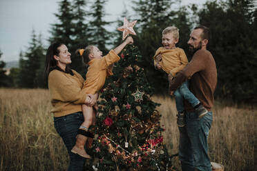 Eltern mit glücklichen Kindern beim Schmücken des Weihnachtsbaums auf dem Lande bei Sonnenuntergang - GMLF00607