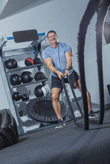 Smiling mature man exercising with battle rope in gym - VTF00629