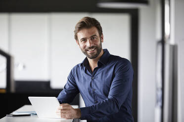 Smiling businessman using digital tablet while standing at desk in office - RBF07890