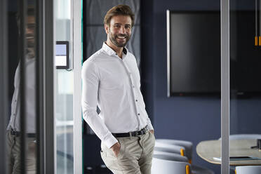 Smiling businessman leaning on glass wall in office - RBF07898