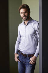 Man with hands in pockets standing against green wall in office - RBF07912