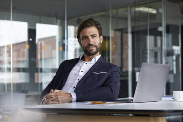 Thoughtful businessman with laptop sitting on chair in office - RBF07921