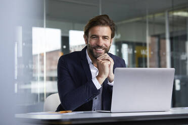 Smiling businessman using laptop while sitting on chair in office - RBF07922