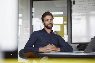 Thoughtful businessman sitting with hands clasped in office - RBF07927