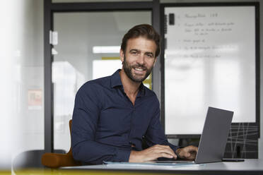 Smiling man working on laptop while sitting by desk in office - RBF07928