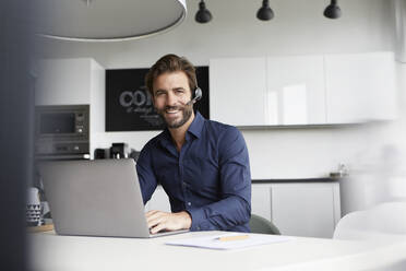 Geschäftsmann mit In-Ear-Kopfhörer im Gespräch bei der Arbeit am Laptop in der Cafeteria - RBF07933