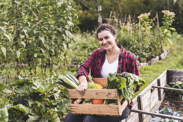 Lächelnde junge Frau mit Gemüse in einer Kiste, die an Pflanzen im Garten lehnt - UUF21432