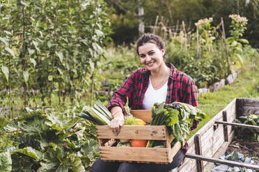 Smiling young woman with vegetables in crate sitting against plants at garden - UUF21432