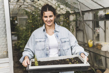 Smiling young woman holding tray while standing in greenhouse - UUF21450