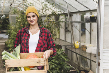 Lächelnde junge Frau trägt Gemüse in einer Kiste, während sie vor einem Gewächshaus steht - UUF21453