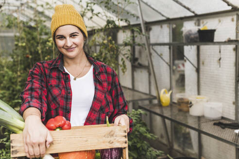 Lächelnde Frau trägt Gemüse in einer Kiste, während sie vor einem Gewächshaus steht - UUF21454