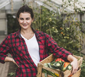 Young woman carrying crate with vegetables while standing against greenhouse - UUF21457