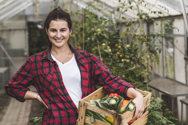 Lächelnde Frau, die eine Gemüsekiste trägt und vor einem Gewächshaus steht - UUF21458