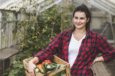 Junge Frau, die eine Kiste mit Gemüse trägt und gegen ein Gewächshaus blickt - UUF21459
