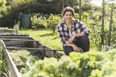 Lächelnde junge Frau, die bei einem Hochbeet im Gemüsegarten hockt - UUF21464