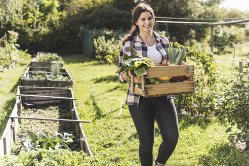 Junge Frau trägt Kiste mit Gemüse beim Spaziergang im Gemeinschaftsgarten - UUF21467