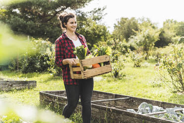 Lächelnde junge Frau trägt Holzkiste mit Gemüse im Gemeinschaftsgarten - UUF21468