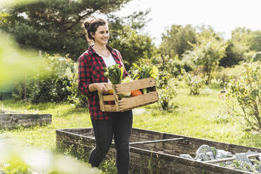 Smiling young woman carrying wooden crate with vegetables in community garden - UUF21468