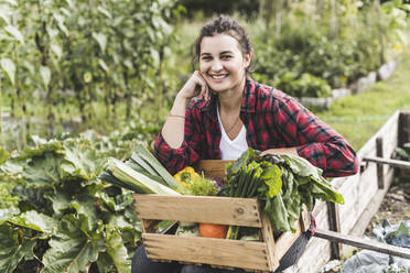 Smiling young woman sitting with vegetables in wooden crate at community garden - UUF21471