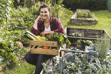 Smiling young woman sitting with vegetables in crate on raised bed at community garden - UUF21472