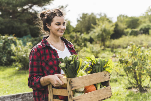 Junge Frau trägt Gemüse in einer Kiste und schaut weg, während sie im Garten arbeitet - UUF21473