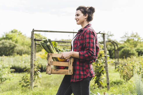 Junge Frau trägt Gemüse in einer Kiste, während sie gegen den Himmel im Garten läuft - UUF21474