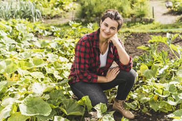 Junge Frau mit Hand am Kinn kniend inmitten von Gemüse, das im Garten wächst - UUF21476