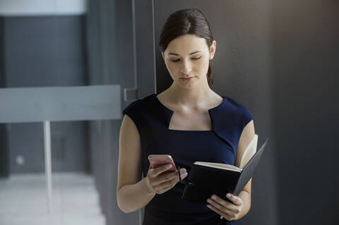 Confident businesswoman holding diary using smart phone while standing against wall stock photo