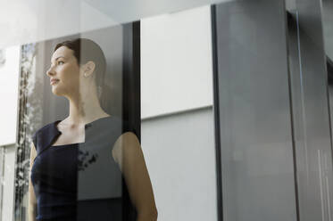 Thoughtful female entrepreneur looking away while standing in office seen through glass door - BMOF00411