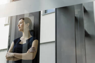 Nachdenkliche Unternehmerin mit verschränkten Armen im Büro stehend, gesehen durch eine Glastür - BMOF00412