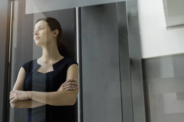 Thoughtful businesswoman with arms crossed standing in office seen through glass door - BMOF00413