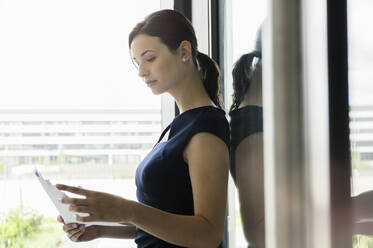 Geschäftsfrau liest Dokumente, während sie am Fenster im Büro steht - BMOF00415
