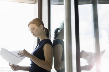 Businesswoman analyzing reports while standing by window in office - BMOF00416