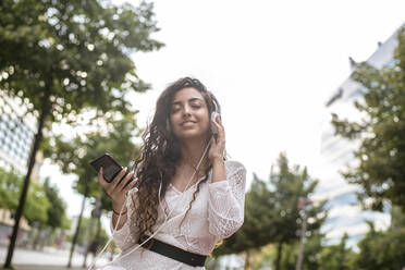 Young woman holding mobile phone with eyes closed listening music against clear sky - BFRF02289