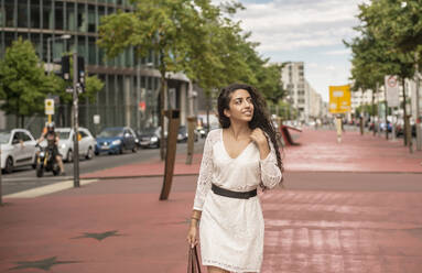 Junge Frau in weißem Kleid schaut weg, während sie auf einem Fußweg in der Stadt geht - BFRF02290