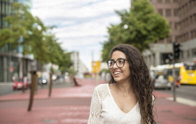 Lächelnde junge Frau mit Brille, die wegschaut, während sie auf einem Fußweg in der Stadt steht - BFRF02291