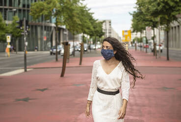 Young woman wearing mask looking away while walking on footpath in city - BFRF02292