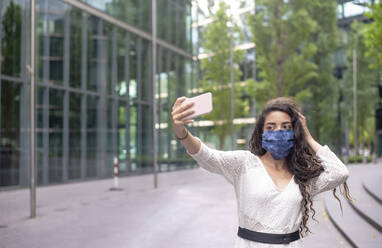 Junge Frau mit Maske, die ein Selfie mit ihrem Smartphone macht, während sie auf einem Fußweg in der Stadt steht - BFRF02295