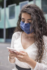 Close-up of young woman wearing face mask using mobile phone while standing in city - BFRF02296