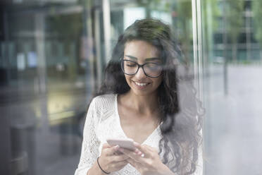 Nahaufnahme einer jungen Frau mit langen Haaren, die ein Mobiltelefon benutzt, während sie am Fenster steht - BFRF02299