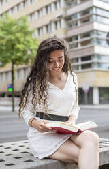 Schöne Frau mit langen Haaren, die ein Buch liest, während sie auf einem Sitz in der Stadt sitzt - BFRF02301