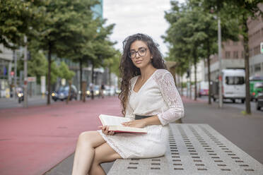 Junge Frau mit langen Haaren liest ein Buch und sitzt auf einem Sitz in der Stadt - BFRF02302