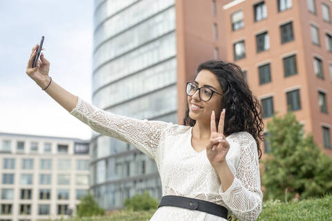 Smiling young woman showing peace sign while taking selfie with smart phone in city stock photo