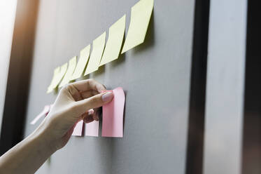 Close-up of businesswoman hand sticking adhesive notes on wall in office - BMOF00421