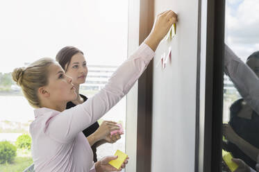 Weibliche Kollegen kleben Haftnotizen an die Wand, während sie in einer Besprechung im Büro diskutieren - BMOF00422