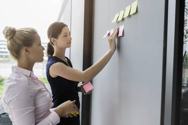 Female colleagues sticking adhesive notes on wall while discussing in office - BMOF00423